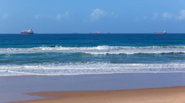 Sahildeki mavi dalgalar kıyı şeridine çarpıyor ve iki gemi liman girişini bekliyor..