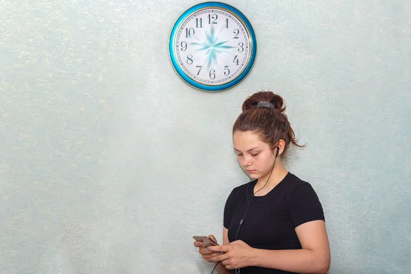 A young girl in headphones stands under the clock and looks at a mobile phone. On the wall clock, the hands seemed to have frozen in motion, time has stopped for the teenager