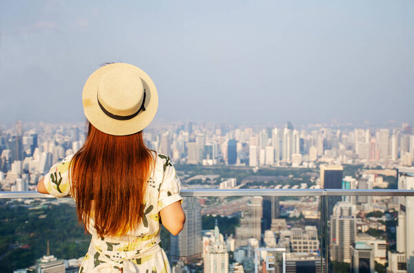 Young woman wear yellow dress and hat, Asian traveler standing and looking aside on balcony with beautiful modern city view on background
