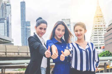 Başarılı genç üç iş kadını mutlu bir şekilde gülümsüyor ve şehrin arka planına başparmak kaldırıyor..