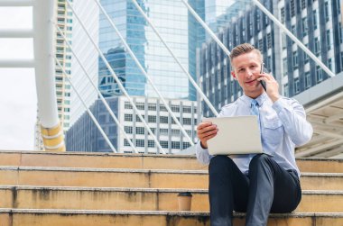 Genç ve yakışıklı bir işadamı dizüstü bilgisayarının yanında oturuyor ve merdivenlerde telefonla konuşurken kahve içiyor.
