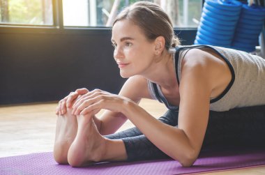 Yoga yapan genç bir kadın, paspasın üzerine oturmuş öne eğiliyor ve bacaklarını tutuyor. Ön tarafta oturan kadın yoga egzersizi yapıyor. Paschimottanasana pozu, spor kıyafeti, seçici odak yüzü.