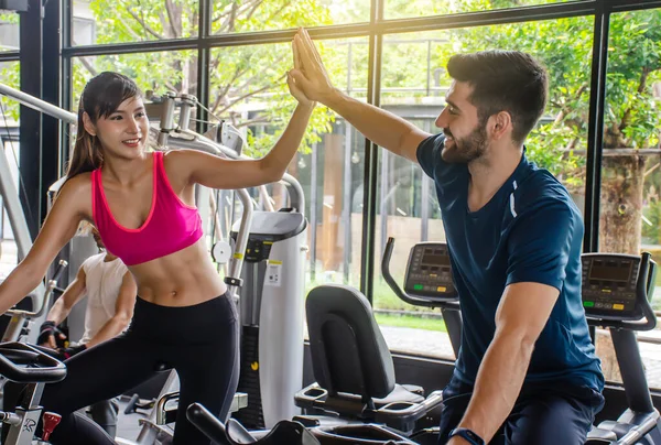 Young sporty woman and man giving each other a high five after cycling training in gym. Fit couple high five after workout in health club, Fitness class doing sport biking in the gym for health.