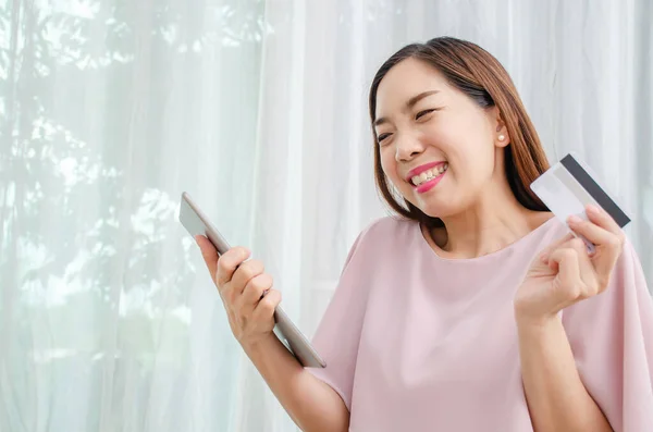 Smiling asian woman holding discount credit card in hand paying for shopping online at tablet in home, Young casual lifestyle