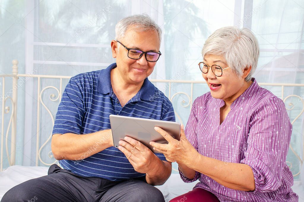 Portrait of smiling senior couple with eyeglasses using electronic tablet while relaxing at the bed room.