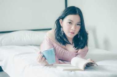 Genç Asyalı kadın kitap okuyor ve sabahları yatakta sıcak kahvenin tadını çıkarıyor.