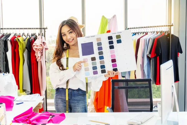 Asian woman designer standing smiling with holding show fabric color sample at the designer desk in the studio. Clothes designers are working in the office. Startup designer concept.