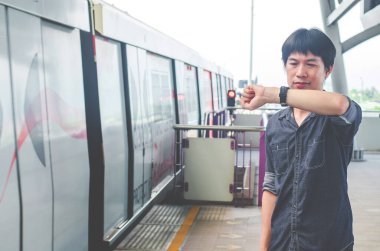 Asyalı bir adamın bir şey beklerken ya da zaman hakkında endişelenirken durup saatine bakması. Arka plan olarak hareket treni. İş ve seyahat kavramı.