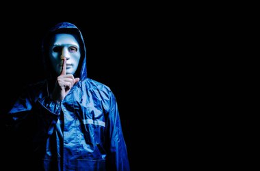 Beyaz maskeli ve kapüşonlu isimsiz bilgisayar korsanı. Karanlık yüz, karanlık arka planda sessizlik jesti yapıyor, veri hırsızı, internet saldırısı, darknet ve siber güvenlik kavramı.