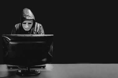 Beyaz maskeli ve kapüşonlu isimsiz bilgisayar korsanı. Karanlık yüz ortada duruyor ve bilgisayar, veri hırsızı, internet saldırısı, darknet ve siber güvenlik konseptiyle bilgisayarı hackliyor..
