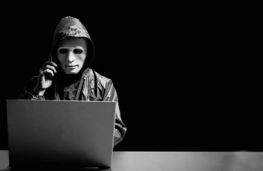 Anonymous computer hacker in white mask and hoodie. Obscured dark face using laptop computer for cyber attack and calling on cellphone, Data thief, internet attack, darknet and cyber security concept. clipart
