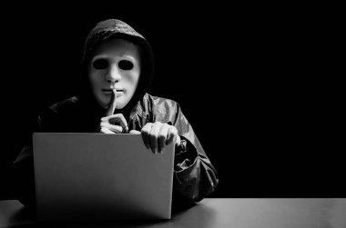 Beyaz maskeli ve kapüşonlu isimsiz bilgisayar korsanı. Karanlık yüz, karanlık arka planda sessizlik jesti yapıyor, veri hırsızı, internet saldırısı, darknet ve siber güvenlik kavramı.