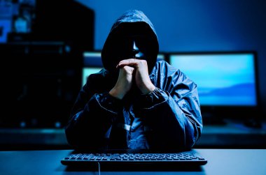 Beyaz maskeli ve kapüşonlu isimsiz bilgisayar korsanı. Karanlıkta klavye bilgisayarı olan karanlık bir yüz. Veri hırsızı, internet saldırısı, darknet ve siber güvenlik kavramı.