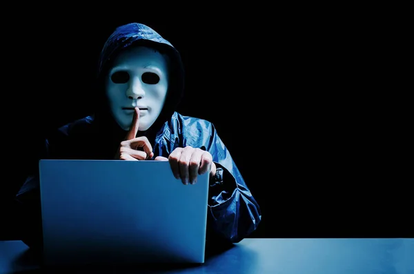 Anonymous computer hacker in white mask and hoodie. Obscured dark face making silence gesture on dark background, Data thief, internet attack, darknet and cyber security concept.
