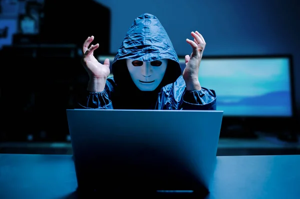 Anonymous computer hacker in white mask and hoodie. Stressful male hacker screaming on a damaged laptop caused by virus, Data thief, internet attack, darknet and cyber security concept.