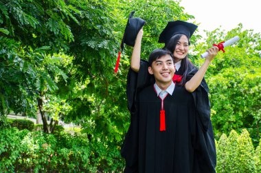 Siyah mezuniyet elbisesi giymiş bir erkek ve kadın ya da mezuniyet şapkasıyla mezun olmuş bir çift ayakta duruyor, ona arkadan sarılıyor, gülümsüyor ve arka planda parkın şapkası var.
