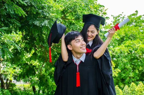 A man and woman couple dressed in black graduation gown or graduates with congratulations with graduation hats is standing, she is hugging him from the back is smiling and holding hat with the park background