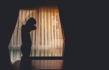 Karanlık odada perdelerle pencerenin önünde duran yalnız adam silueti. Adam tek başına pencerede duruyor. Depresyon ve endişe bozukluğu kavramı..