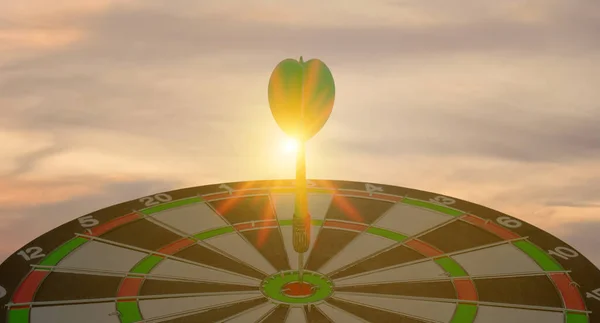 Silhouette of green dart arrow hitting bullseye target center dartboard on sunset background. Business targeting and focus concept, strategy, achievement, and planning concept