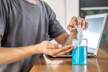 Ofiste dizüstü bilgisayarla çalışmaya başlamadan önce el dezenfektanı kullanan Asyalı adam, mikropları, bakterileri ve virüsü öldürmek için pompa şişesinde temizleyici.