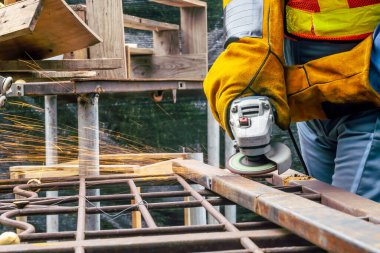 Çalışan mavi üniformalı genç bir kaynakçı ve çalışan eldivenler garajdaki metal bir objeyi öğütüyor. Parlak kıvılcımlar yanlara doğru uçuyor.