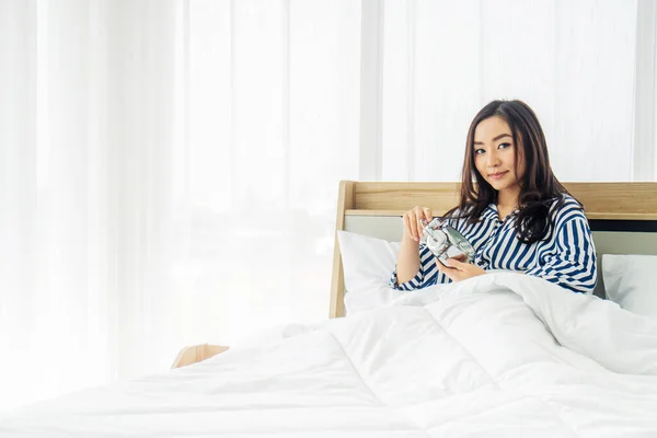 Asian beautiful woman wake up on bed  is holding clock to turn off  alarm clock at 7 am in her bedroom. Set up alarm to get up to go working in weekday.