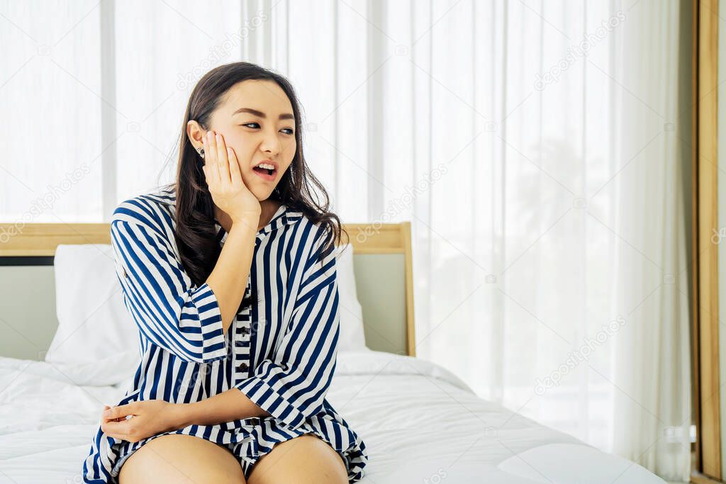 Young Asian woman touching cheek while suffering from toothache and discomfort on bed in white bedroom morning.Concept of women's health care.