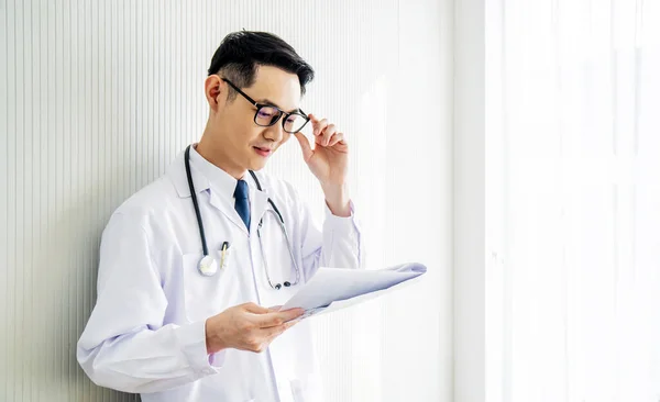Young  asian doctor man wearing lab coat looking down reading to his patient notes standing in hospital