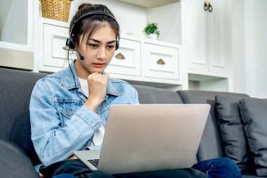Asyalı genç iş kadını kendi evindeki kanepede oturuyor kulaklık takıyor bilgisayarla video konferans yapıyor ve ev ofisinden yeni bir proje düşünüyor, evden çalışıyor.