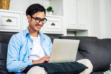 Evdeki bir kanepede günlük kıyafetlerle oturan zeki Asyalı işadamı, bilgisayar bilgisayarını kullanarak online işten ve evden para kazanarak işten zevk alıyor.