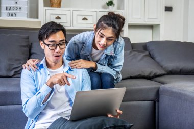 Genç çift koltukta oturmuş bilgisayardan alışveriş yaparak rahatlıyor. Adam ürünü bilgisayar ekranına doğrultuyor..