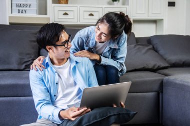 Asyalı çift gülümsüyor ve birbirlerine mutlu bir şekilde sohbet ediyorlar. Evlerinde online alışveriş yapıyorlar, evlerinin rahatlığında kanepede oturuyorlar, rahatlıyorlar ve kolayca mal alıyorlar.
