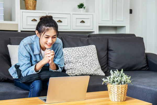 Asian teenage girl sitting on a sofa in the house is enjoying the work on the computer laptop, chatting through video conferencing with friends, work from home