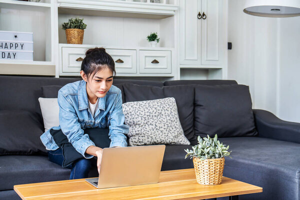 Asian teenage woman sitting on a sofa in the house is enjoying the work by using the computer laptop to make money from online business, work from home
