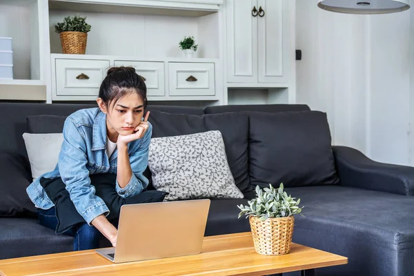 Asian teenage woman sitting on the sofa at home is stressed about her work on her computer laptop,  work from home