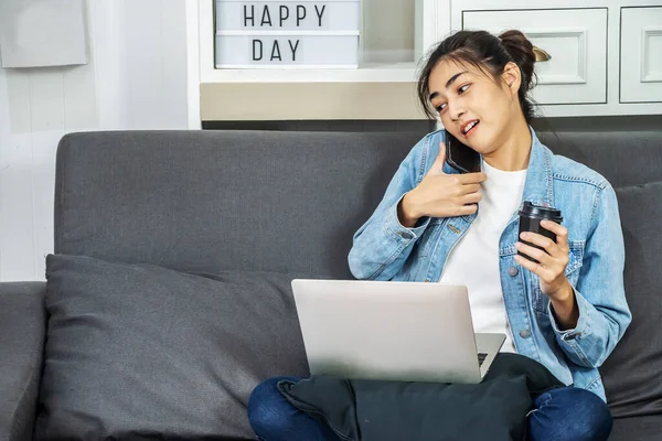Asian woman is sitting on the sofa in her own house is drinking coffee with a phone happy talking to colleagues while working on the computer laptop, work from home