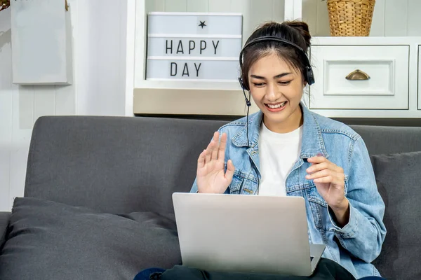 Asian young business woman is sitting on the sofa in her own house is wearing headphones video call conference with laptop computer raise hand saying hi team at home office, work from home