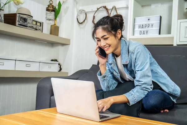 Asian young business woman is sitting on the sofa in her own house is talking on cell phone and smiling with laptop computer at home office, work from home