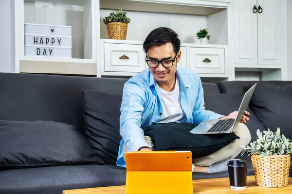 Smart Asian businessman in casual clothing sitting on a sofa in the house is working on computer laptop and tablet , SME start up concept, work from home