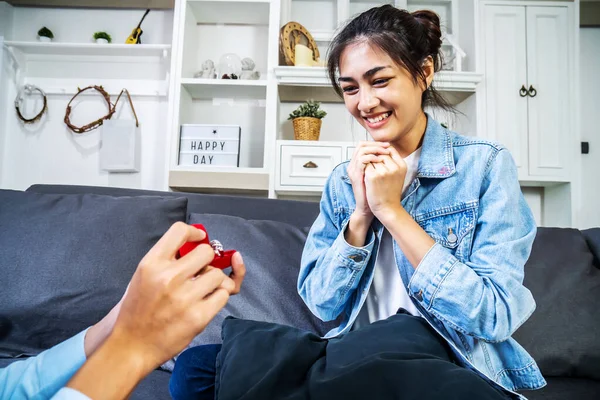Will you marry me? Young man giving engagement ring in little red box to his beautiful and happy girlfriend at home. Man making marriage proposal to his beloved woman. Romantic date.