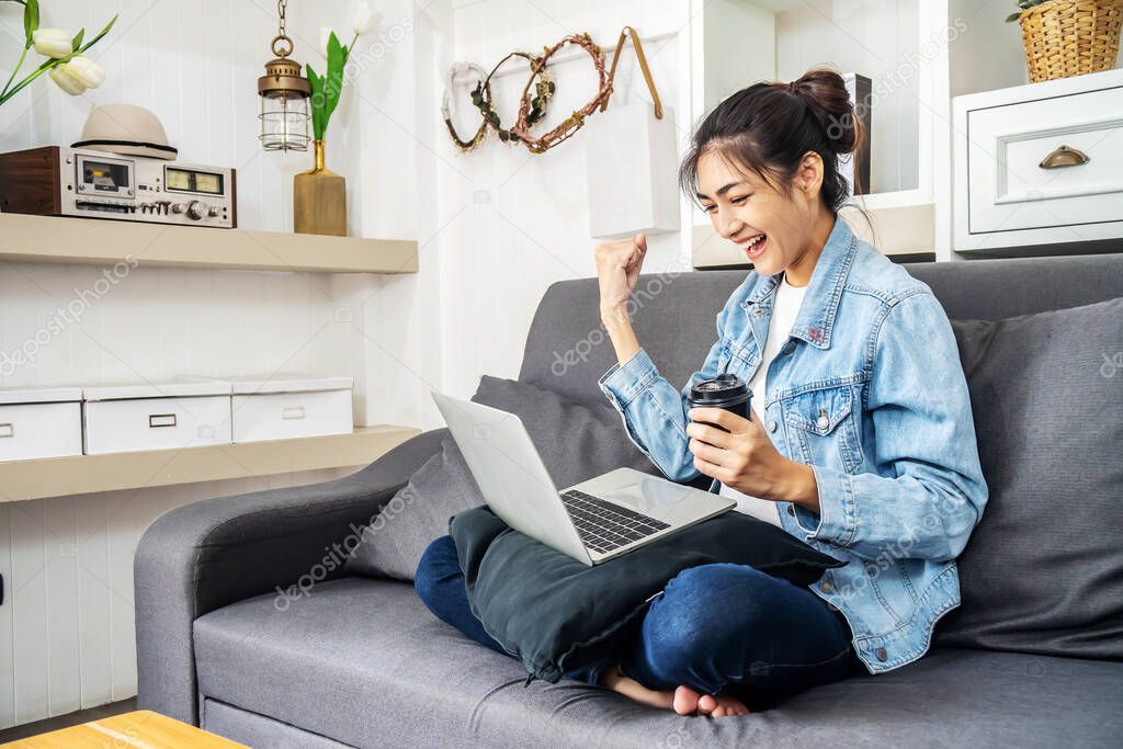 Asian teenage woman sitting on a sofa in the house drinking coffee is enjoying the work by using the computer laptop to make money from online business, work from home