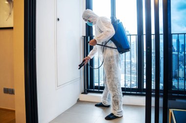 Temizlik için COVID-19, yüz kalkanı, maske ve alkol jeli kullanan ve balkonda, karantinada, dezenfeksiyonda kimyasal dezenfekte kullanan koruyucu koruyucu giysili Asyalı adam veya PPE giysisi