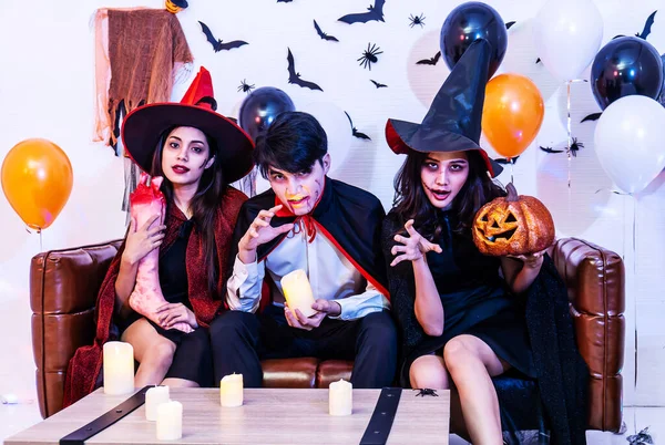 Asian Happy friends sitting on sofa in costumes and makeup on a celebration of Halloween posing with pumpkin and fake human leg in party, Group of young people enjoying Halloween party with many items at home or nightclub.