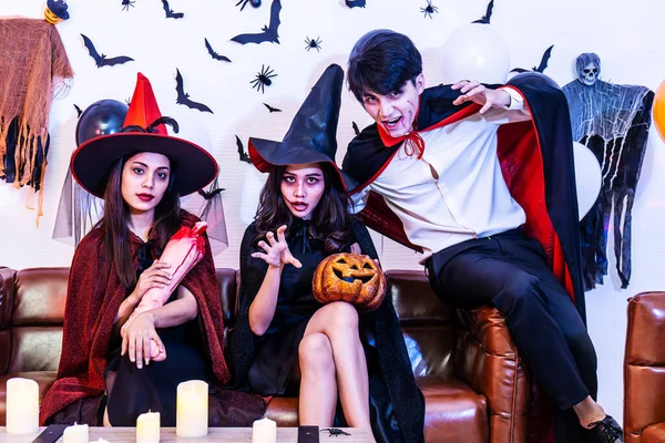 Asian Happy friends sitting on sofa in costumes and makeup on a celebration of Halloween posing with pumpkin and fake human leg in party, Group of young people enjoying Halloween party with many items at home or nightclub.