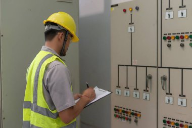 Asyalı elektrik mühendisi kontrol odasındaki elektrik sistemini kontrol etmek ve izlemek için pano tutuyor.