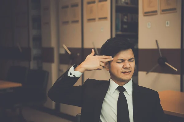 Asian businessman in black suit think idea for work at office,he work overtime alone in a company,dark tone,thailand people,make hand shape gun point on head