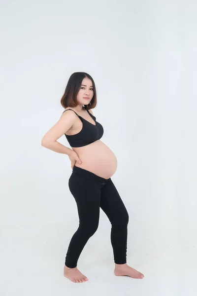 Asian pregnant woman in black undershirt sitting on white bed and