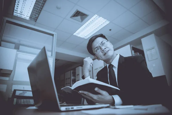 Asian businessman in black suit think idea for work at office,he work overtime alone in a company,dark tone,thailand people