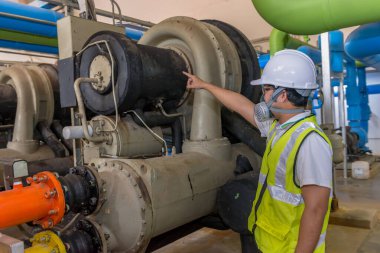 Kazan dairesinde çalışan gözlüklü Asyalı mühendis, ısıtma sistemi ekipmanlarının teknik verilerini kontrol ediyor, Taylandlılar gaz maskesi takıyor.