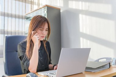 Asyalı kadın telefonda konuşuyor ve ofiste dizüstü bilgisayarla çalışıyor.